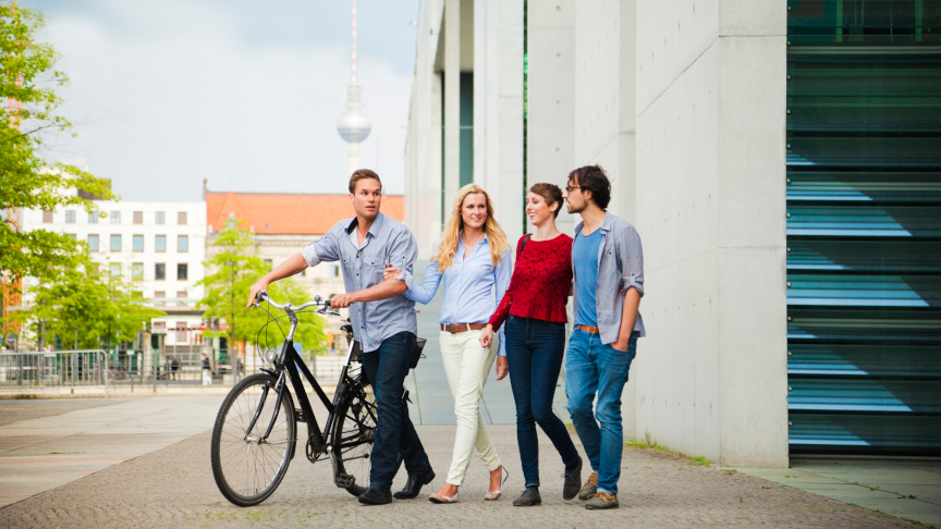 Four students walk through Berlin on a bicycle, the TV tower can be seen in the background.